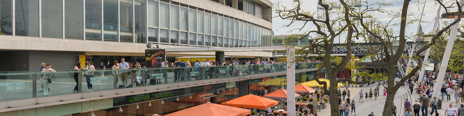 Crowds of people are milling around outside the Southbank Centre in a hive of activity; a view of orange square market stall canopies stretches into the distance. Trees on the right, the London Eye and Big Ben in the far distance.